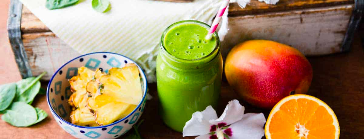 green smoothie in a jar and next to it is pineapple, mango, a halved orange, and a flower