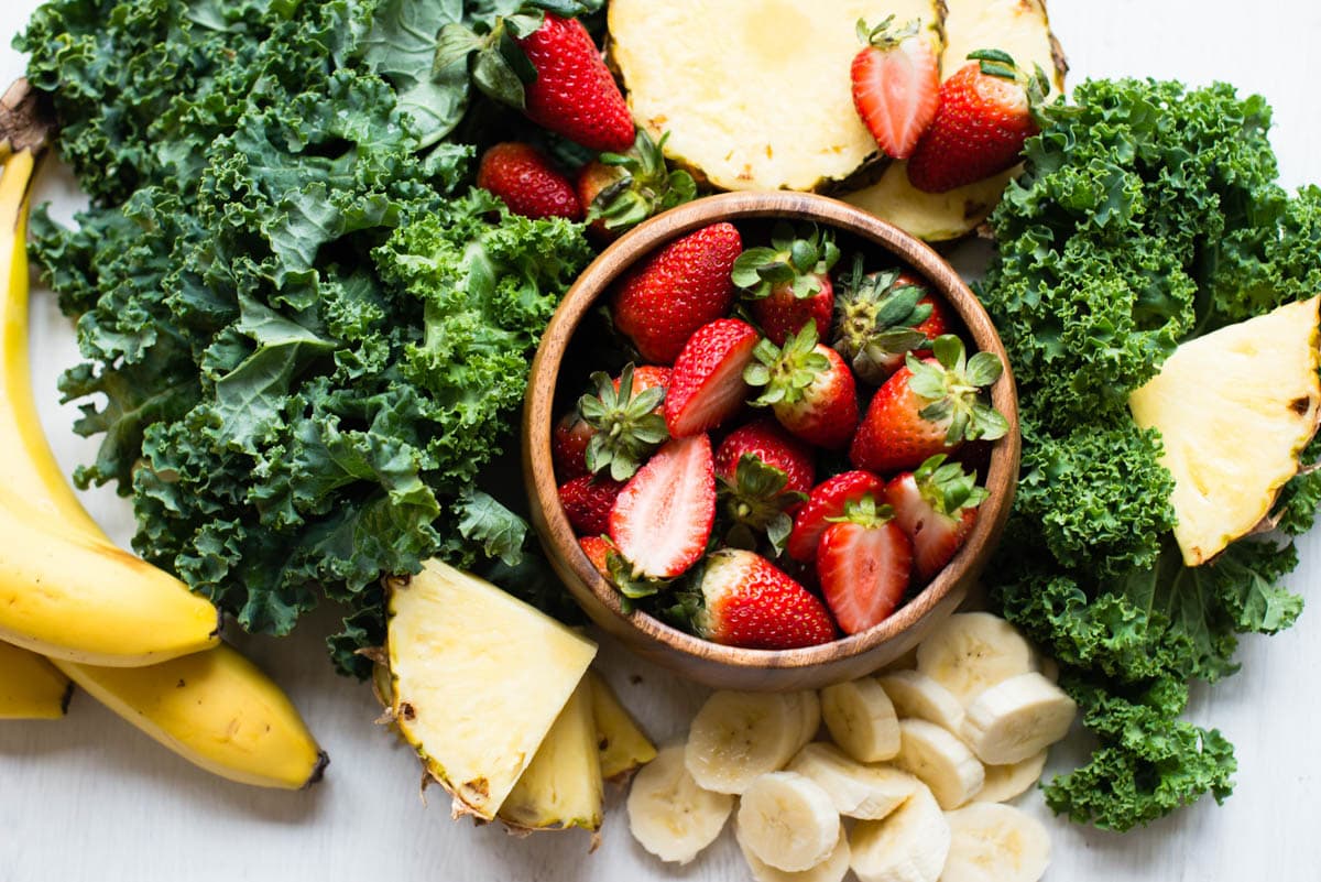fresh ingredients including curly kale, sliced pineapple, bananas and a coconut bowl full of strawberries.