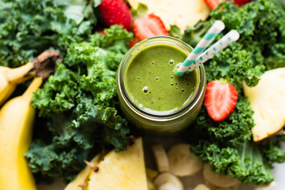 kale banana smoothie in a glass cup surrounded by fresh kale, strawberries, bananas and pineapple.