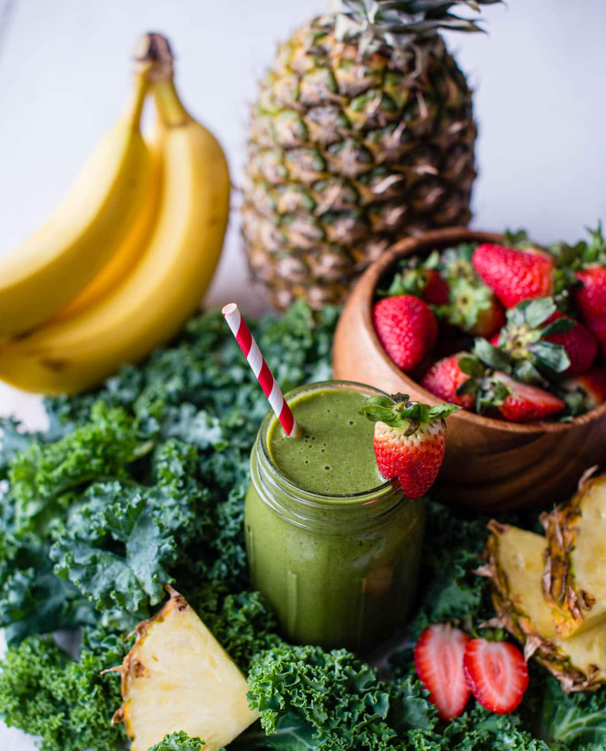 healthy green smoothie in glass jar with fresh strawberry garnish, surrounded by fresh produce.