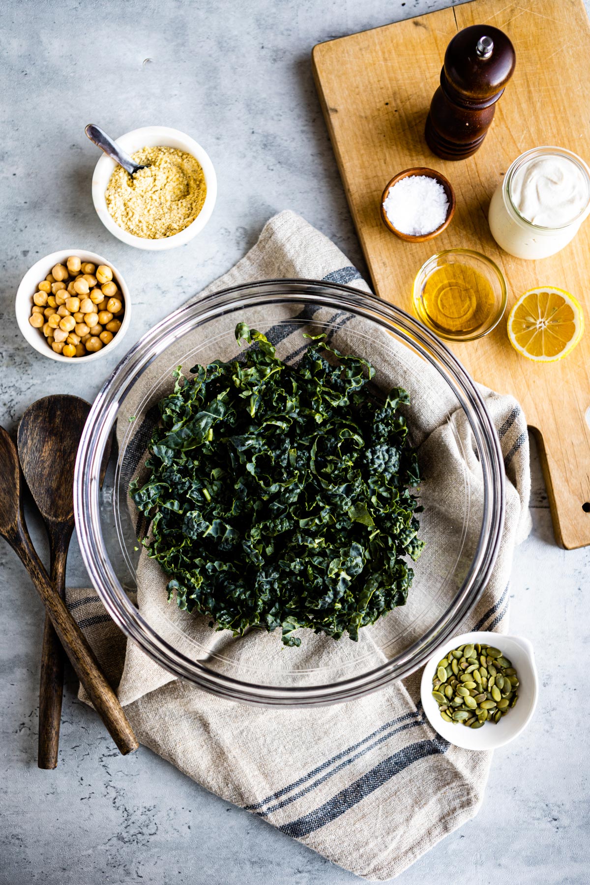 bed of shredded kale in glass bowl next to a small white bowls of pumpkin seeds,. vegan parmesan, chickpeas, garlic aioli, sea salt and olive oil.