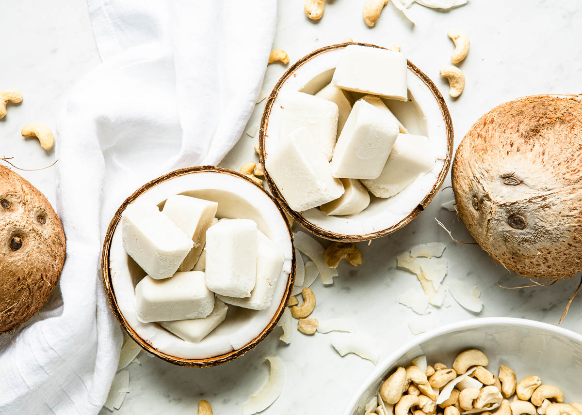 2 coconut halves filled with coconut cubes on a counter with more coconuts, coconut flakes and cashews.