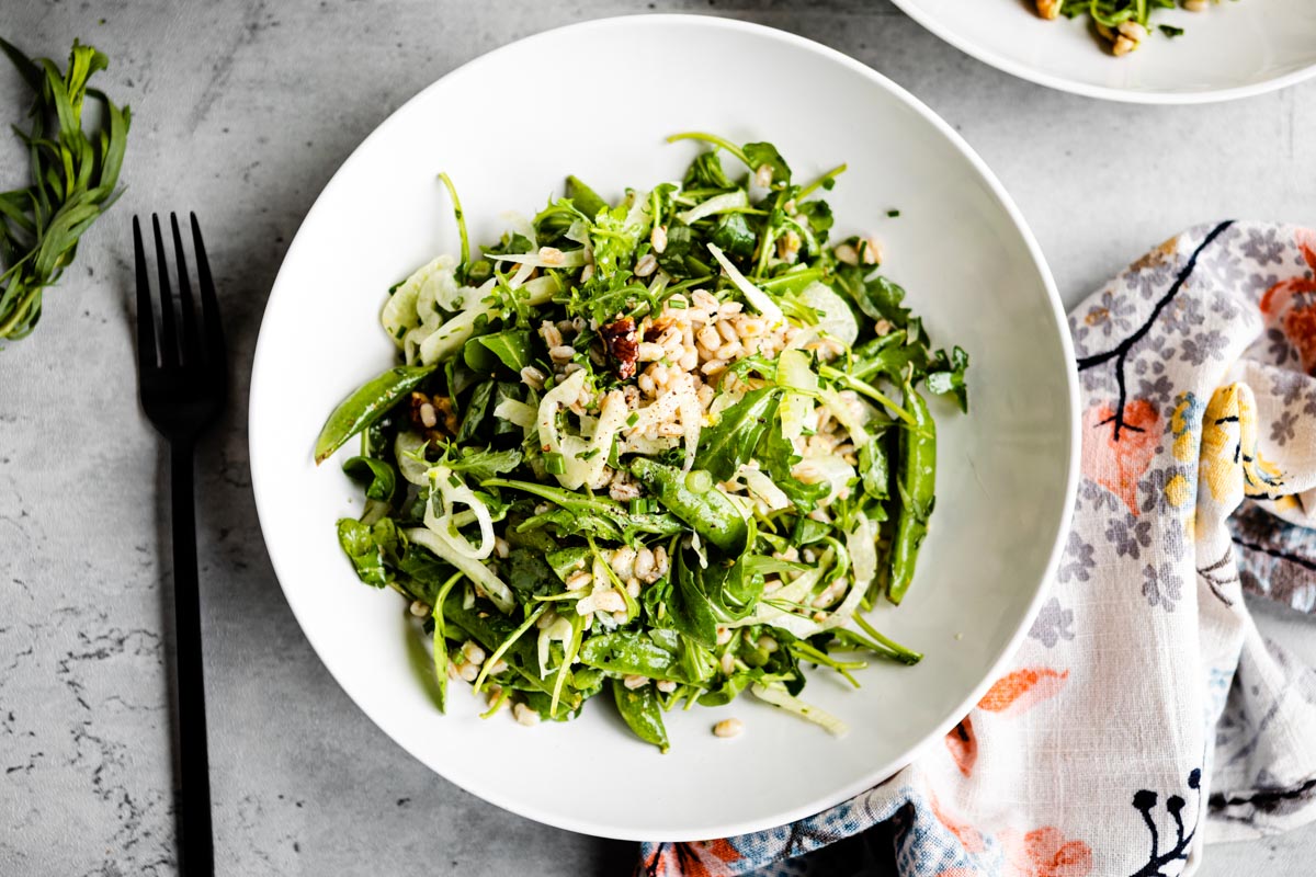 green salad in white bowl topped with grains, walnuts and dressing.