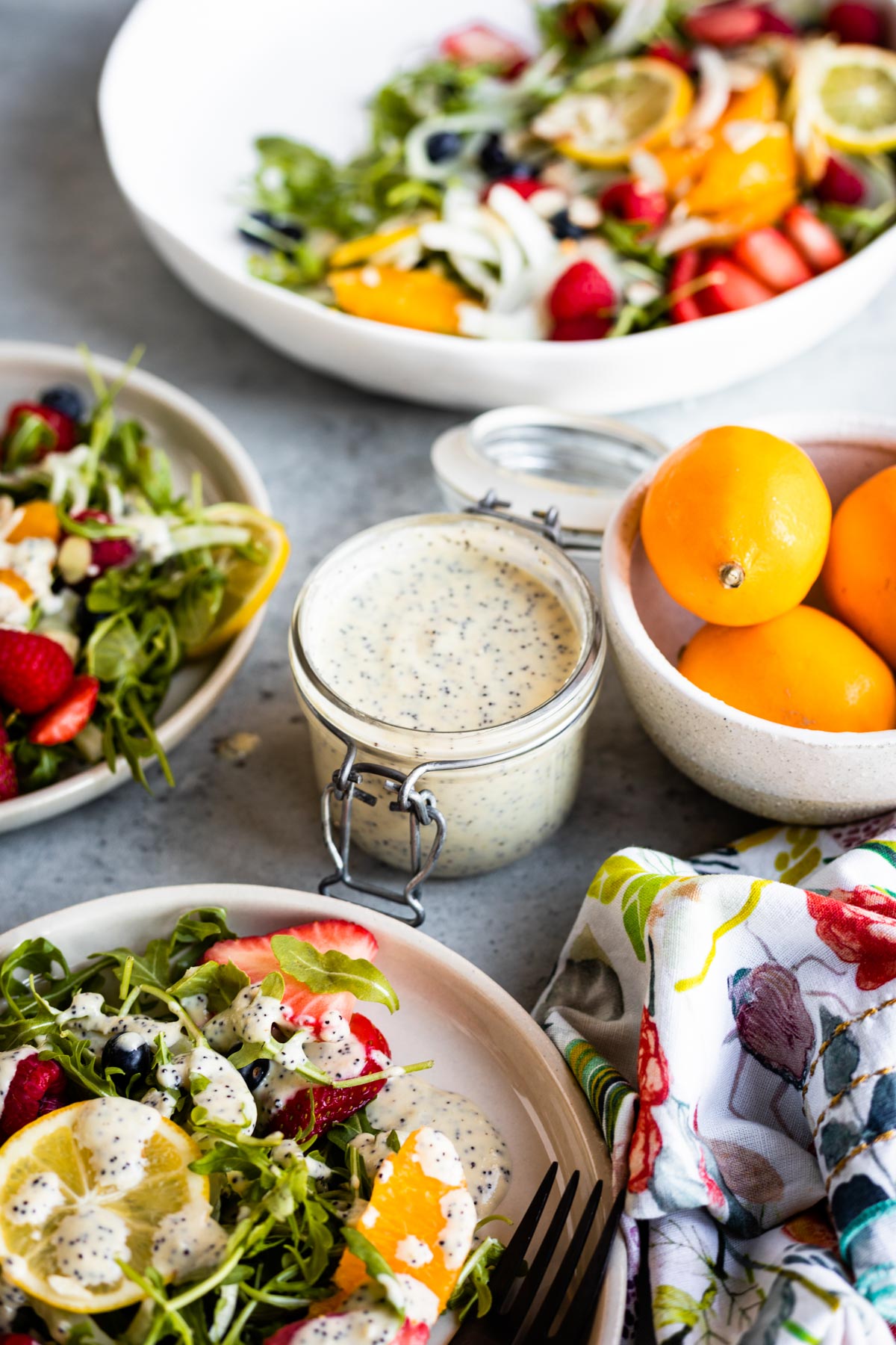 homemade dressing with poppy seeds on top of a fruit and arugula salad as well as in a glass jar next to a bowl of meyer lemons.