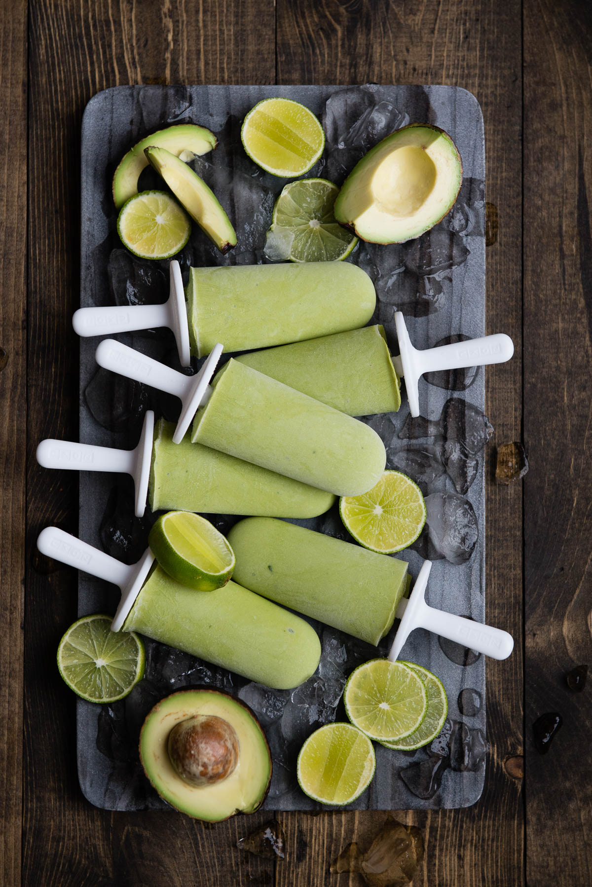 6 lime popsicles on white sticks on a stone slab with ice cubes and lime slices.