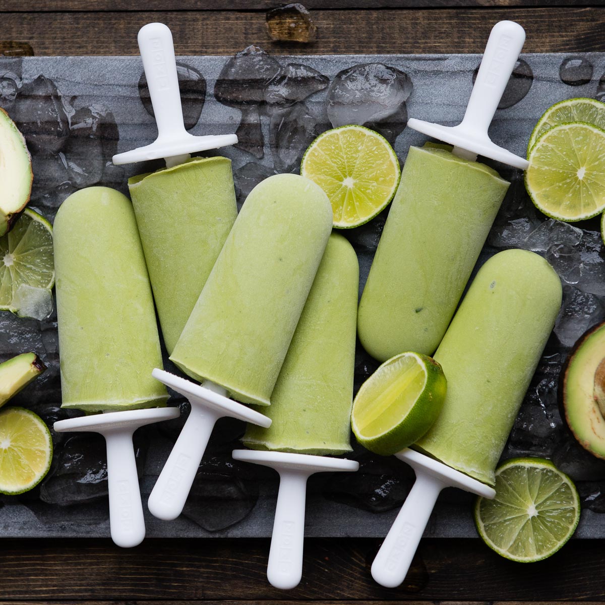 6 lime popsicles on white sticks on a stone slab with ice cubes and lime slices.