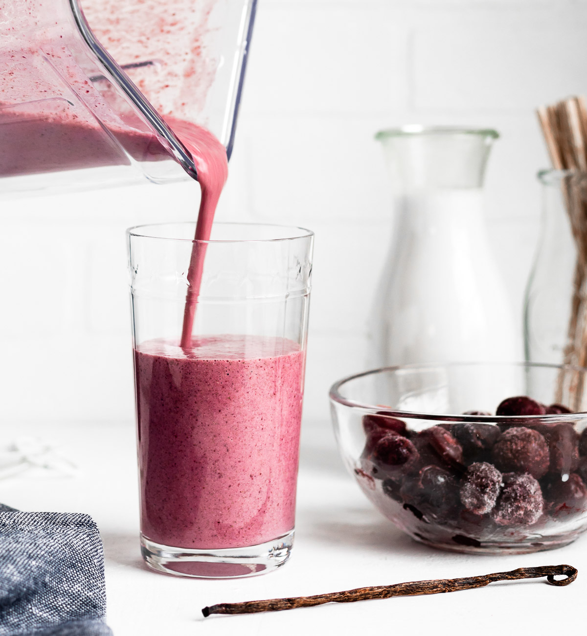 A pink smoothie being poured from a blender into a glass, with frozen cherries and a vanilla bean nearby.