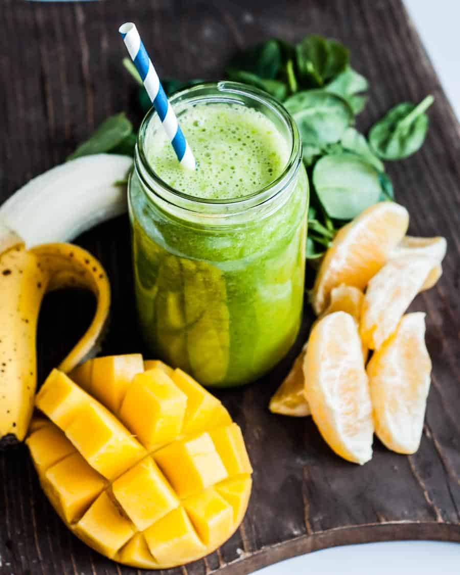 green smoothie in glass jar surrounded by orange slices, spinach, mango and banana.