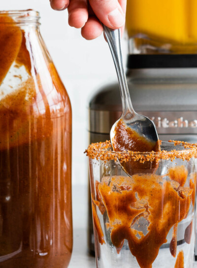 homemade chile suce in a large glass jar and being dripped into a glass with a spoon.