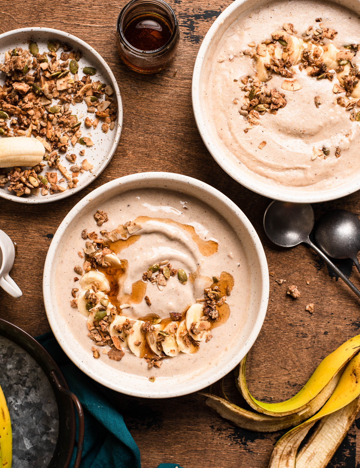 2 oat smoothie bowls topped with maple syrup, granola and sliced bananas, on a wooden counter top.