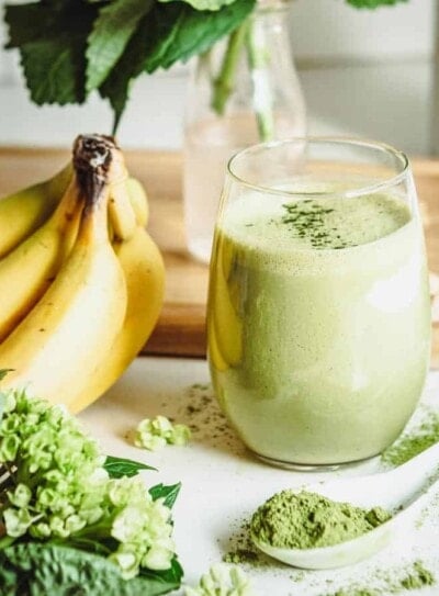 matcha smoothie in a round glass on a white counter top surrounded by fresh ingredients.