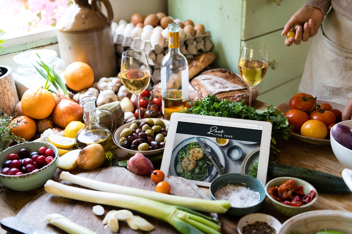 wooden countertop full of ingredients like tomatoes, leeks, olives and oranges and a white table with the words Rawk the Year at the top.