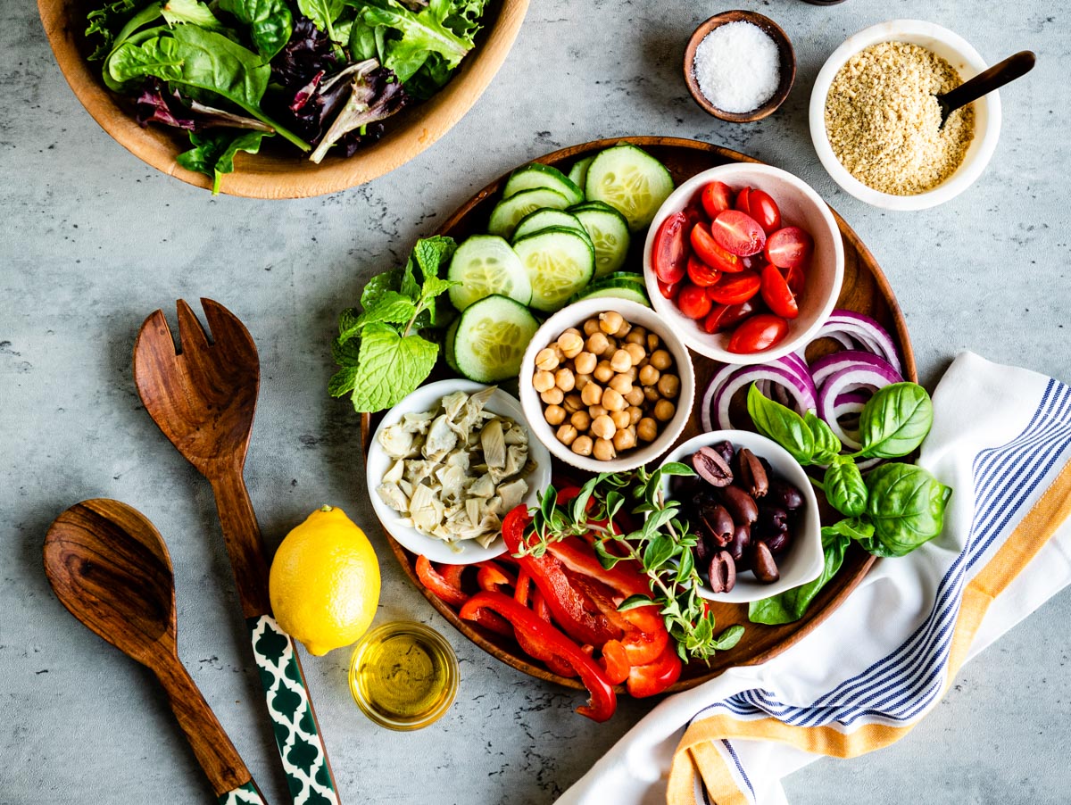 Mediterranean-inspired salad ingredients on a wooden platter next to salad tongs.