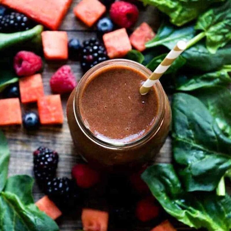 Dark smoothie on a table with fruits and greens.