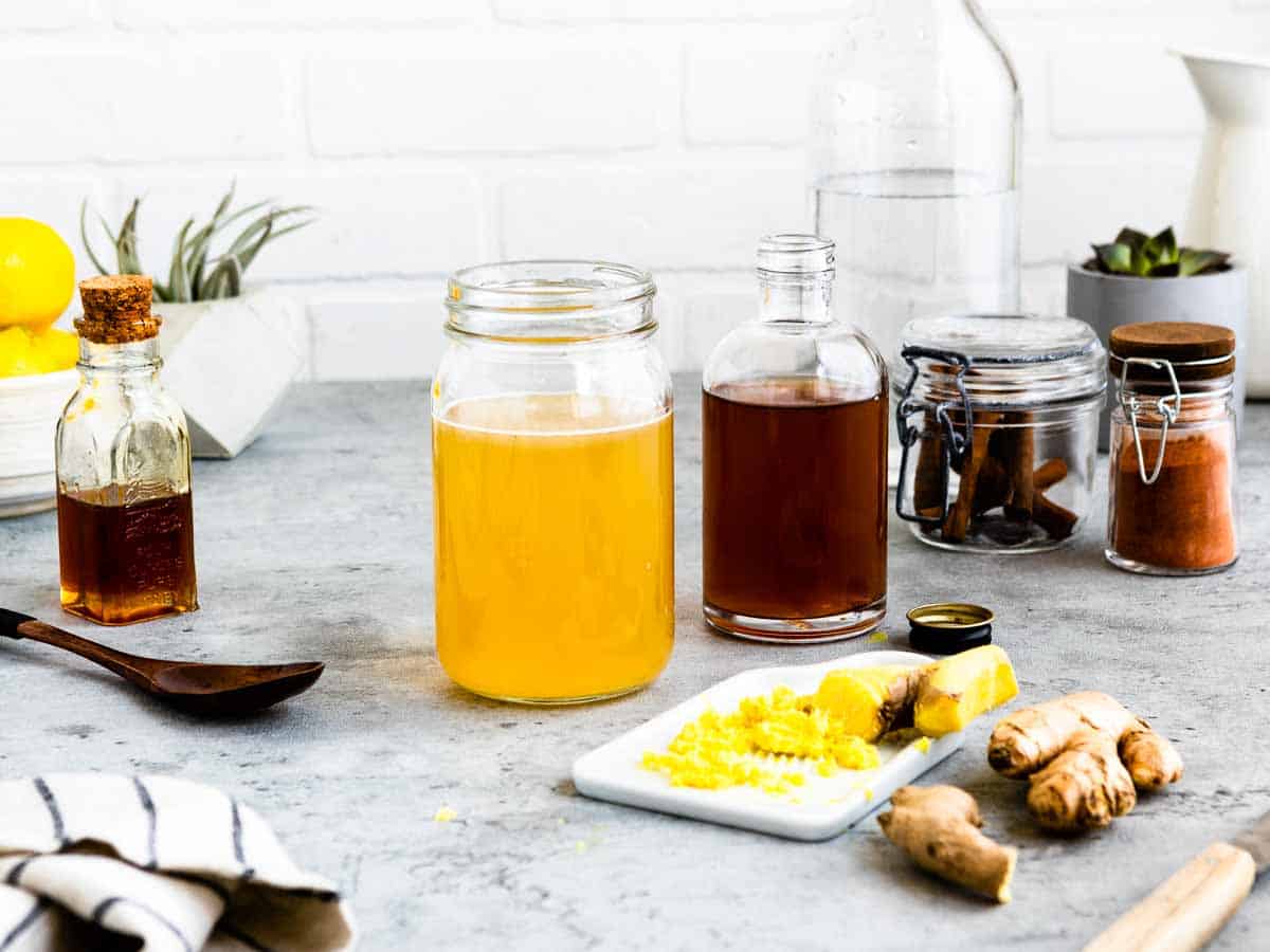 gray counter top with several glass jars of maple syrup, metabolism boosting team, apple cider vinegar and cinnamon as well as fresh grated ginger on a white tray.