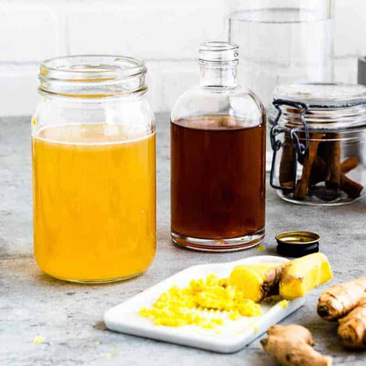 yellow colored tea and apple cider vinegar in glass jars with grated ginger in front on a plate.