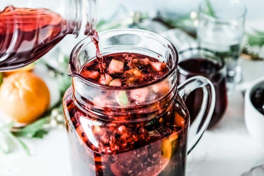 juice being poured into a glass pitcher full of red liquid and fresh sliced fruit.