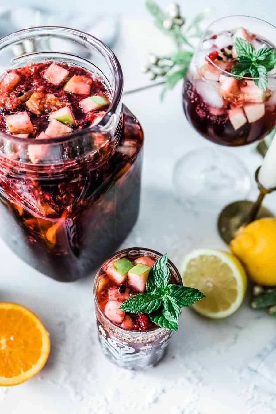 non alcoholic berry sangria loaded with fruit in a glass with mint leaf garnish next to a pitcher of sangria.