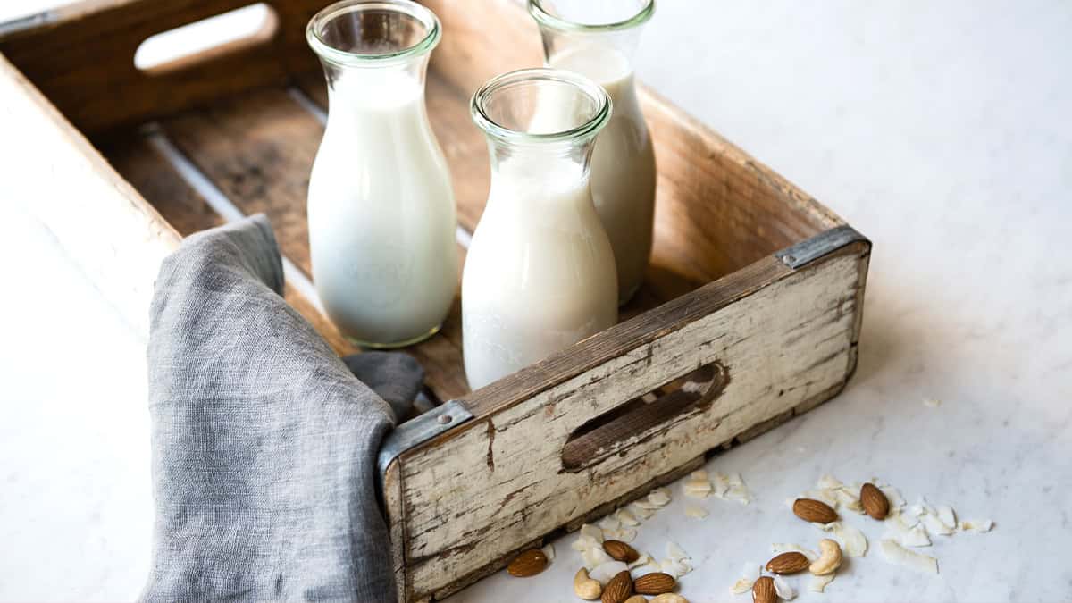 Three glass bottles of plant-based non-dairy milk in a rustic wooden tray.