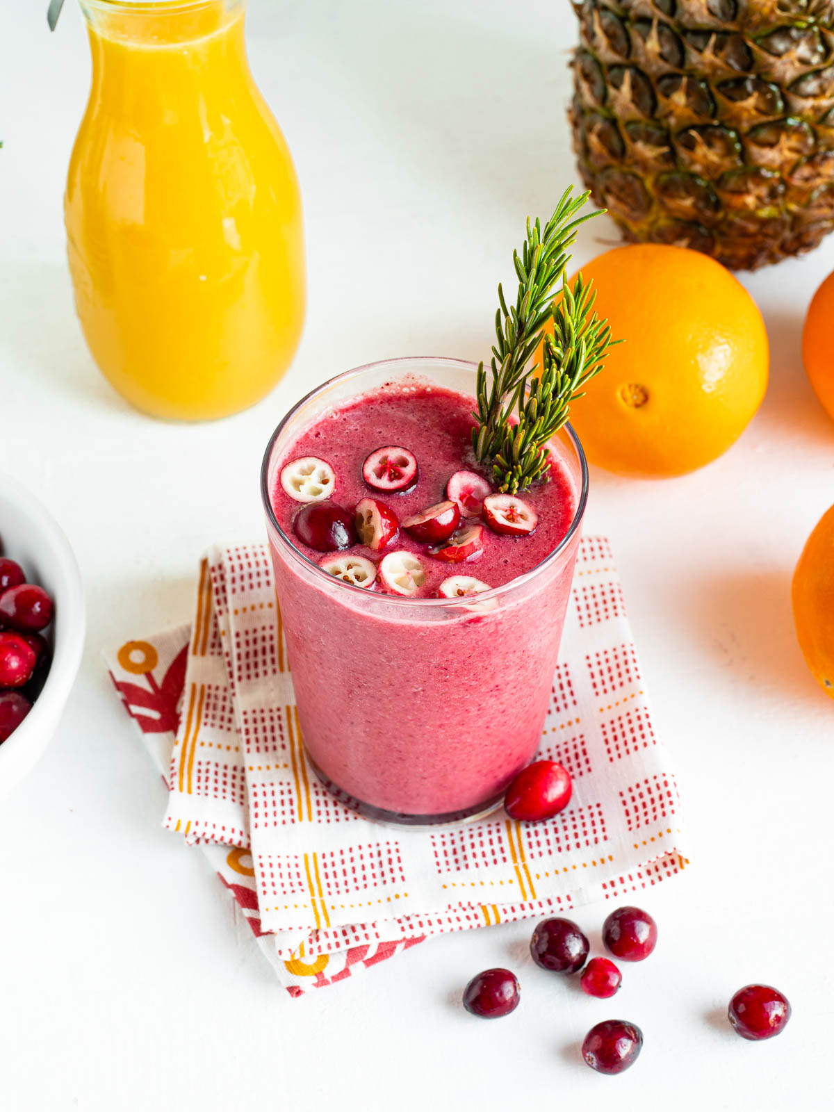 glass full of pink cranberry smoothie topped with fresh sliced cranberries and a sprig of rosemary, sitting on a tea towel. 