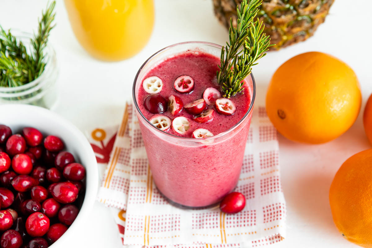 glass full of pink cranberry smoothie topped with fresh sliced cranberries and a sprig of rosemary, sitting on a tea towel. 