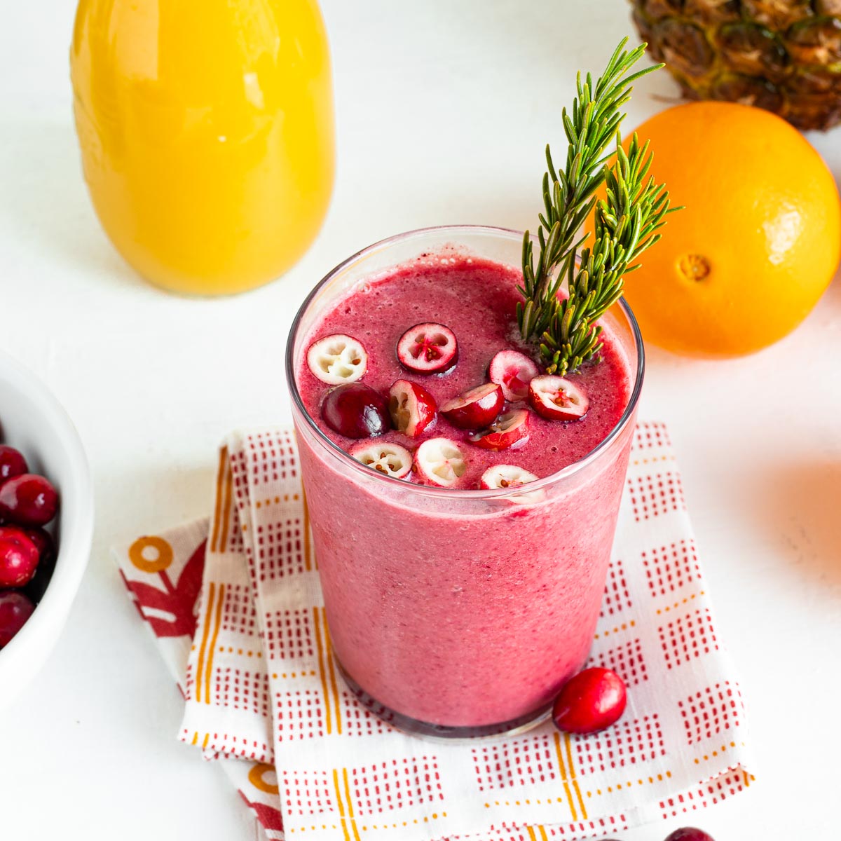 glass full of pink cranberry smoothie topped with fresh sliced cranberries and a sprig of rosemary, sitting on a tea towel.