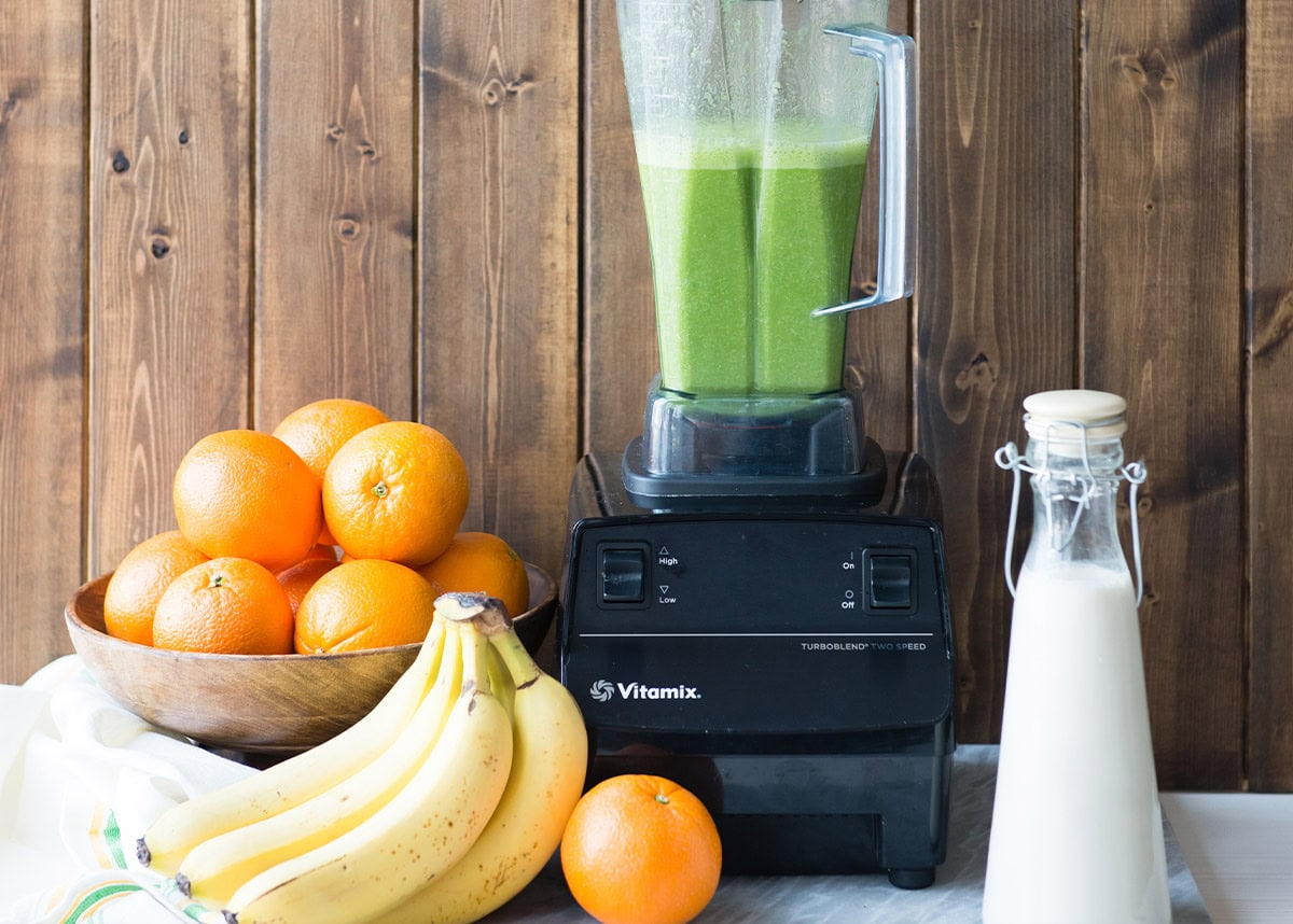 Fresh oranges, bananas, and a green smoothie in a blender on wooden background.