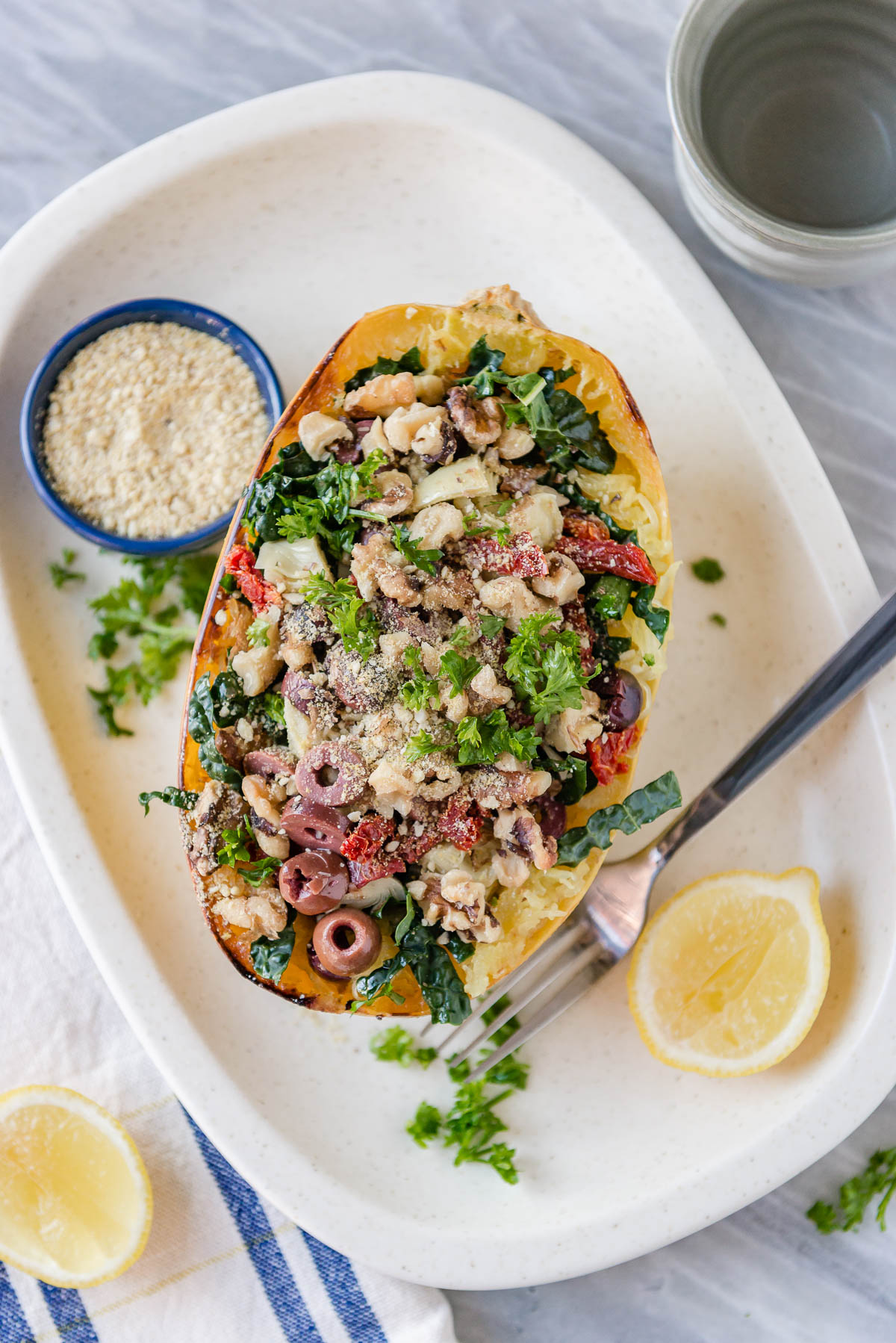 spaghetti squash topped with olives, vegan parmesan, kale and artichokes on a white platter with sliced lemons.