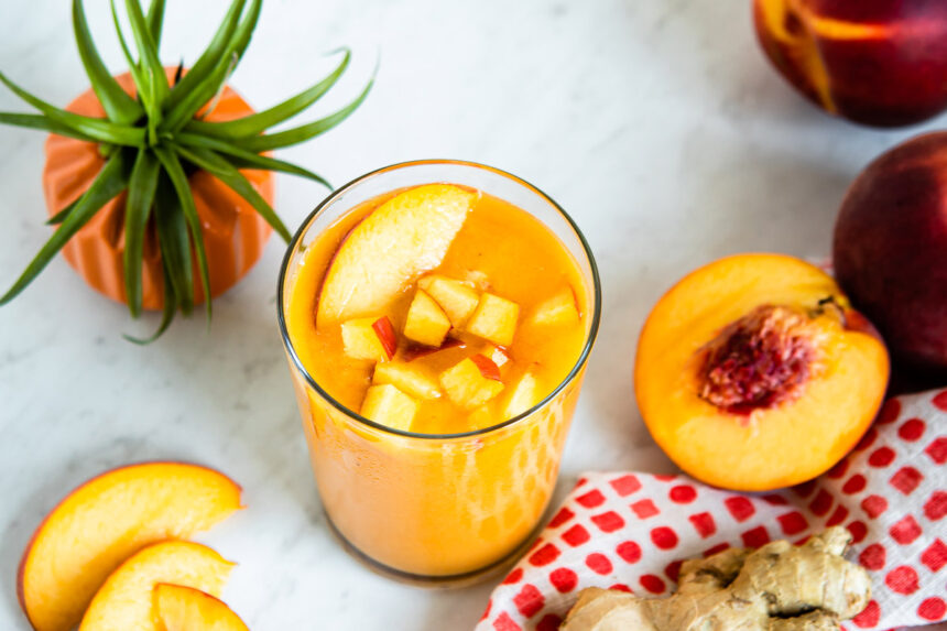 Glass of peach smoothie on white counter with chopped fruit on top.