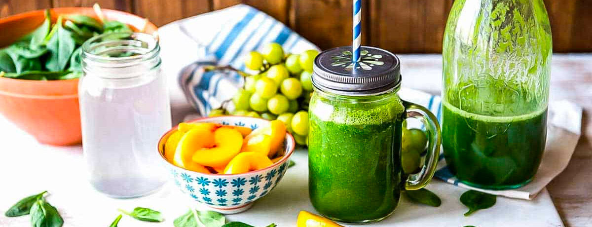 green smoothie with a metal lid and straw with fresh peaches, grapes, and spinach sitting next to it