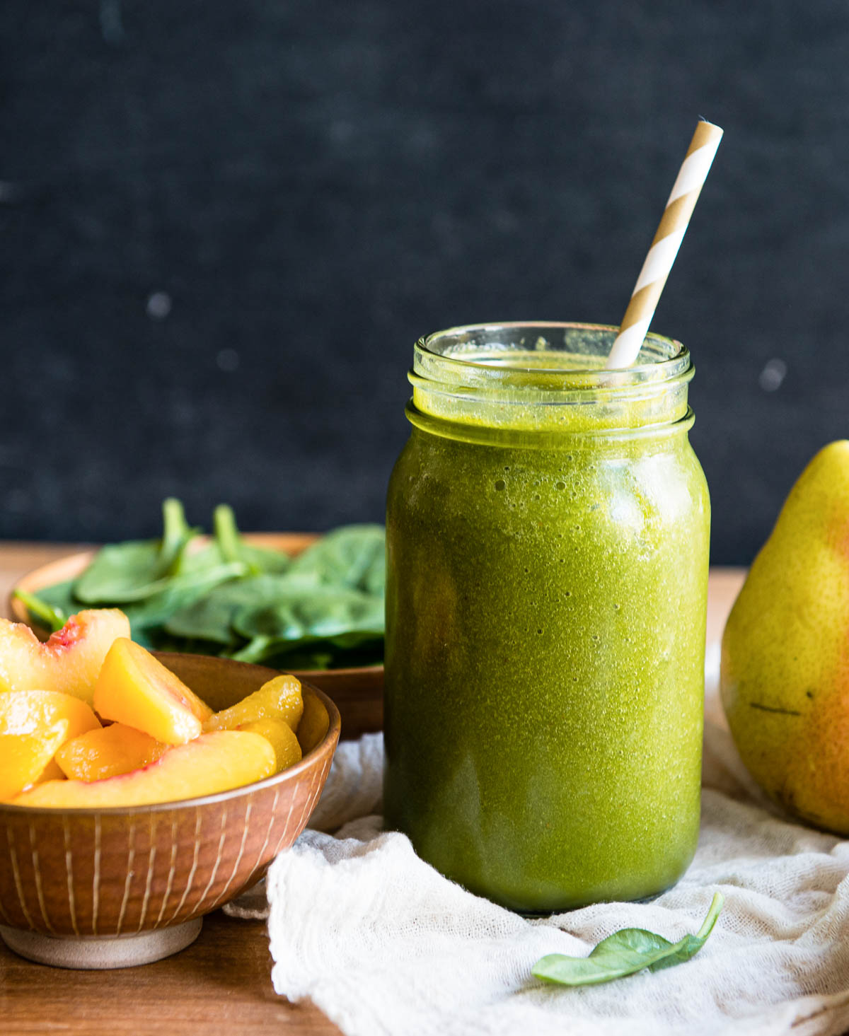 large glass jar with green smoothie and a paper straw, next to peach slices.