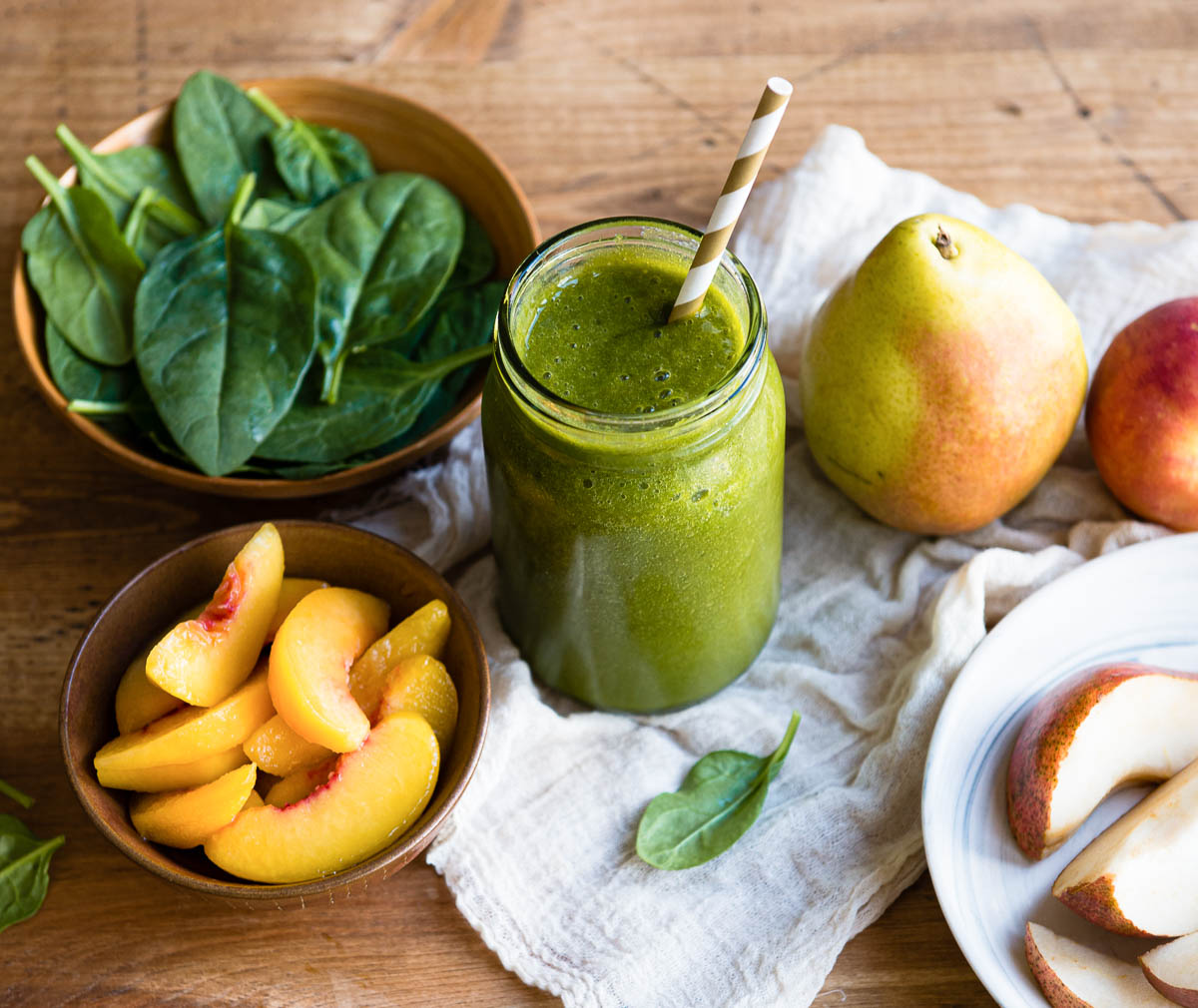 glass jar of green smoothie with a striped paper straw next to pears, spinach and peach slices.