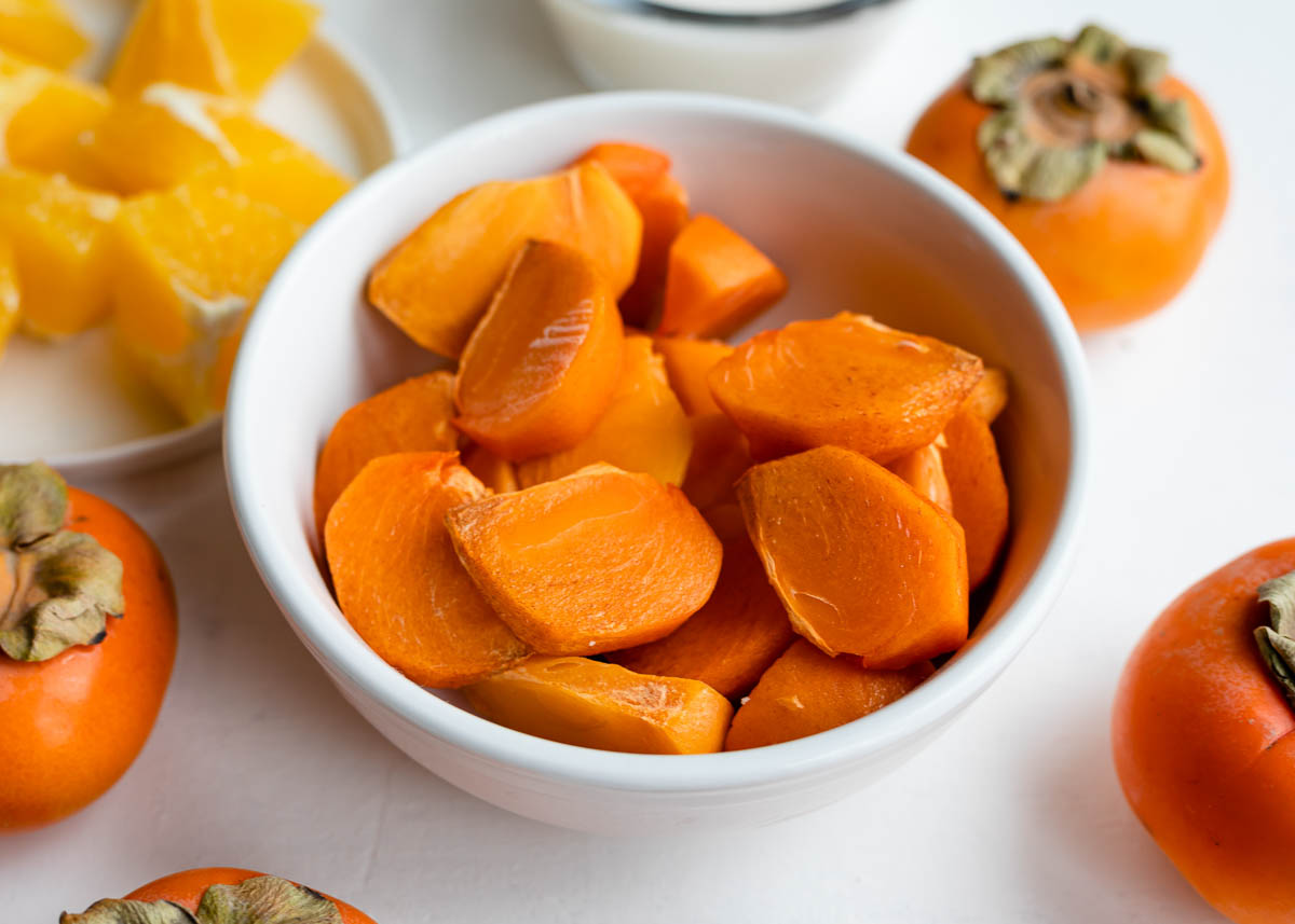 white bowl of persimmon slices on a white countertop next to whole persimmons.
