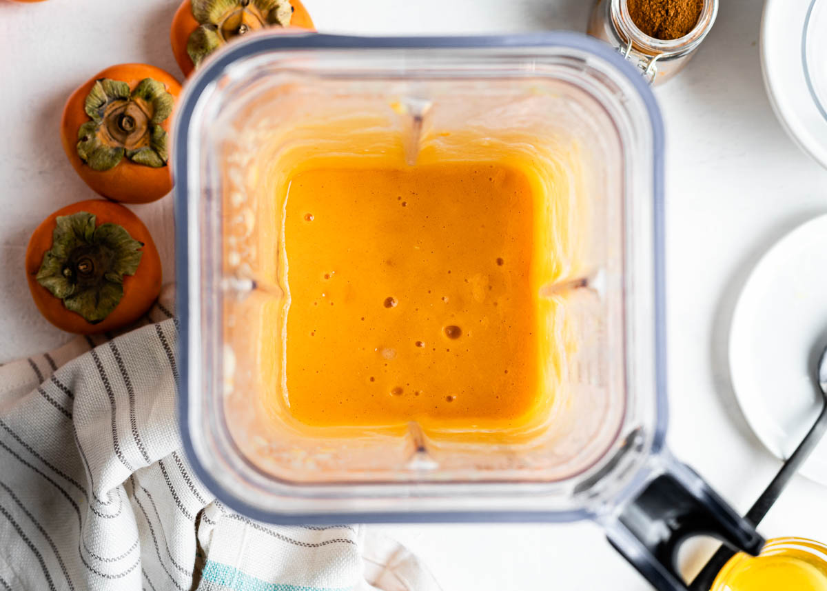 blender container of orange smoothie sitting on a white countertop surrounded by fresh persimmons, a tea towel and a jar of ground cinnamon.