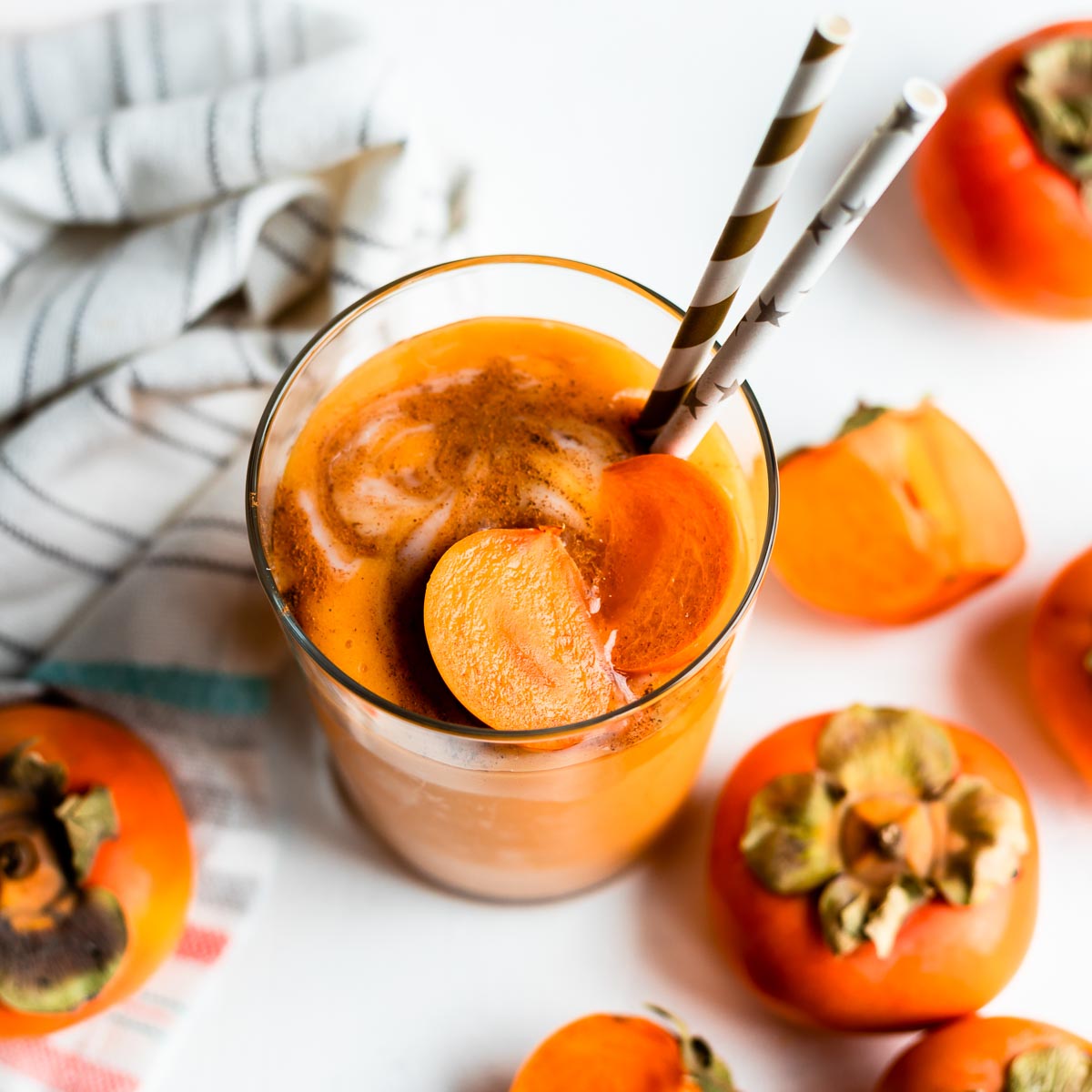 glass of yogurt persimmon smoothie topped with fresh persimmons and 2 paper straws, on a white counter surrounded by persimmons.