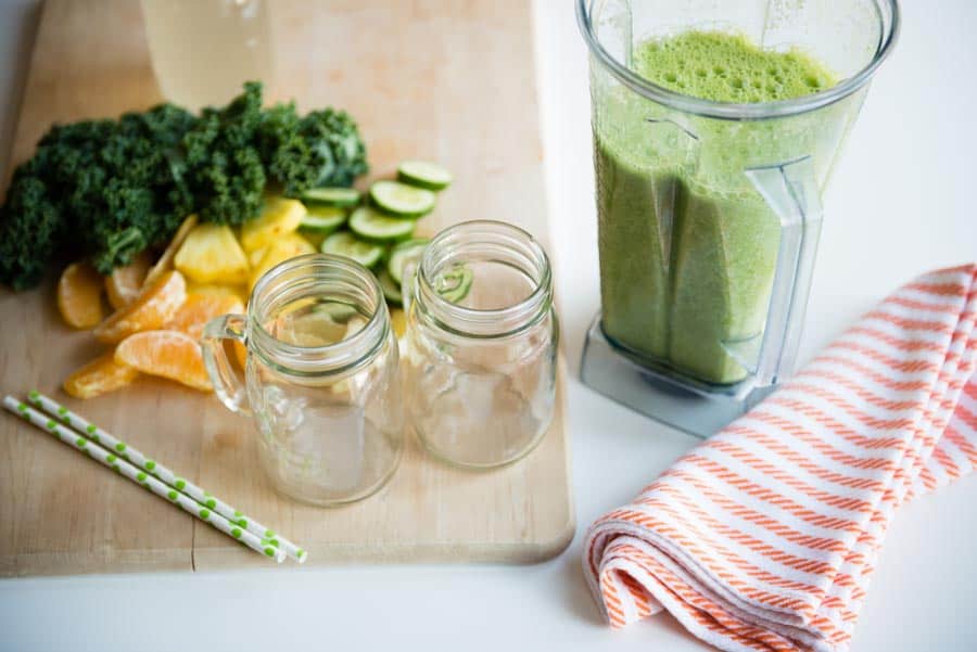 smoothie container full of green smoothie next to 2 empty glass jars, sliced ingredients and paper straws.