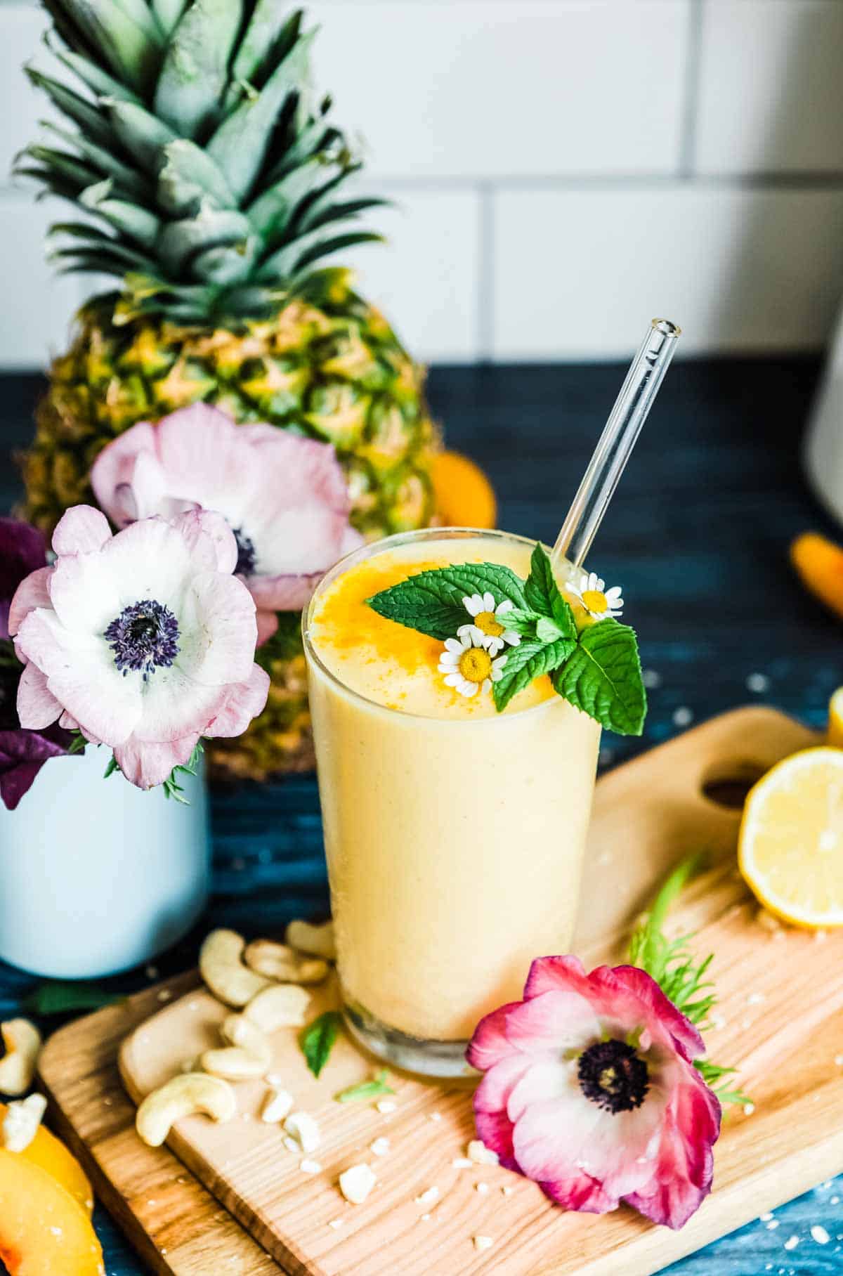 Pineapple smoothie topped with mint and flowers in a glass with a glass straw on a counter top, along with cutting board, ingredients and flowers.