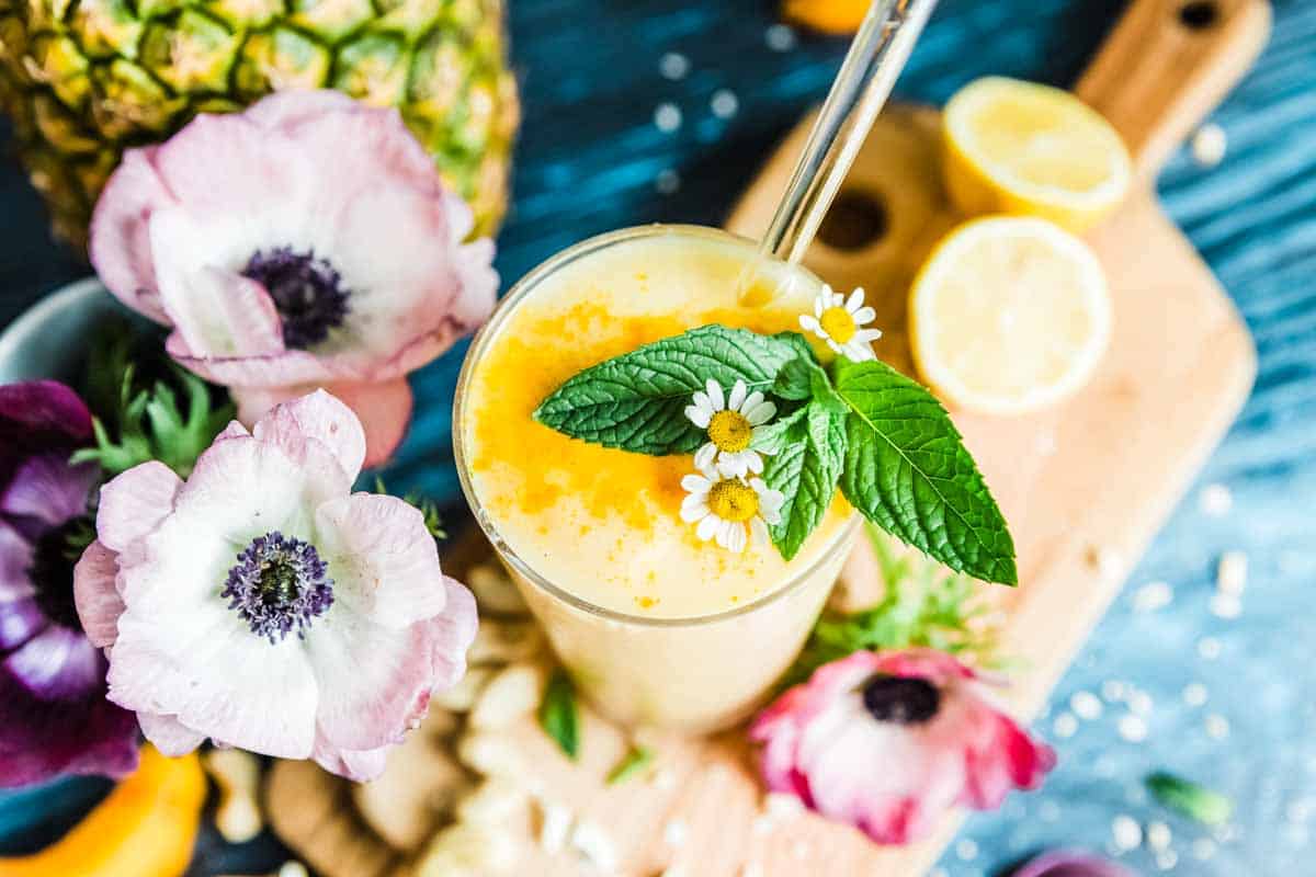 Pineapple juice smoothie in a tall glass with a glass straw and fresh mint leaves, sitting on a wooden cutting board and surrounded by ingredients and flowers. 