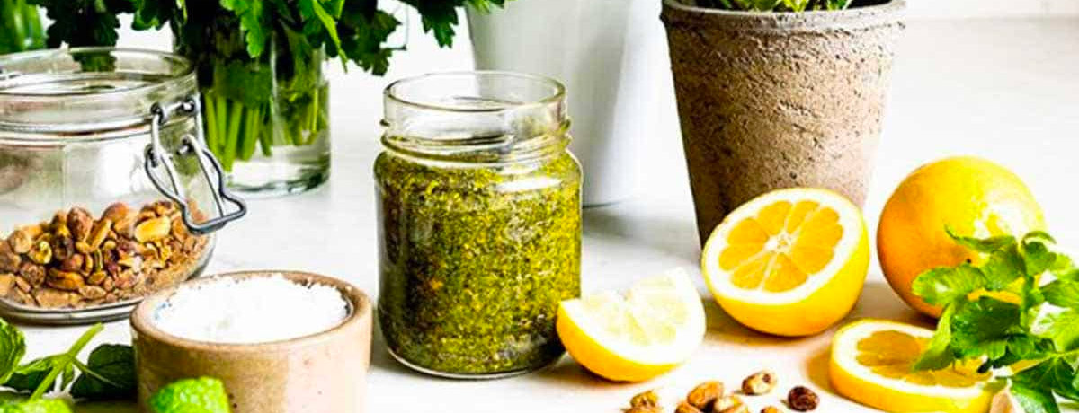 jar of pistachio pesto with ingredients used to make it sitting all around: lemons, pistachios, salt, herbs