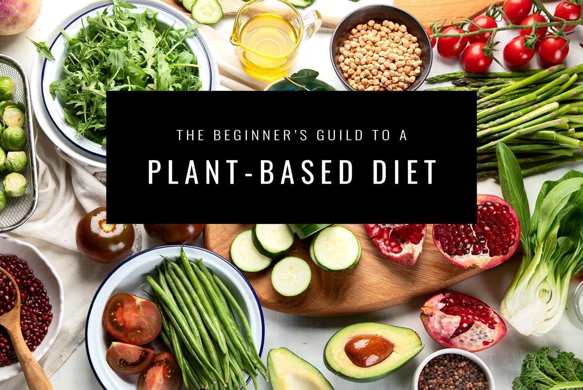 white countertop full of fresh tomatoes, pomegranate, asparagus, bok choy, sliced cucumbers, green beans, arugula, and a black box with white text saying The beginner's guide to a plant-based diet.