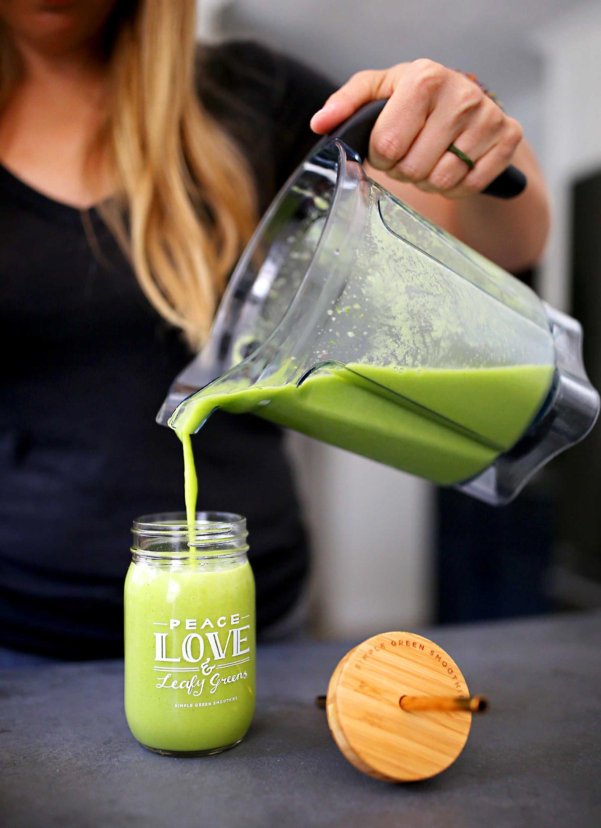A large blender container full of green smoothie being poured into a glass jar with the words Peace Love & Leafy Greens written on it next to a wooden lid and metal straw.