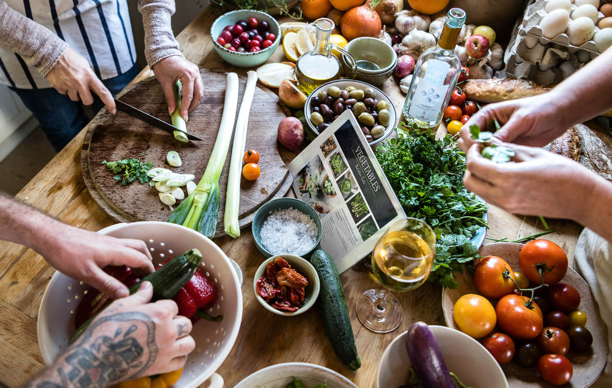 several hands prepping fresh ingredients on a wooden countertop covered in produce like leeks, tomatoes, sun dried tomatoes, olives and an iPade with the word Vegetables on it.