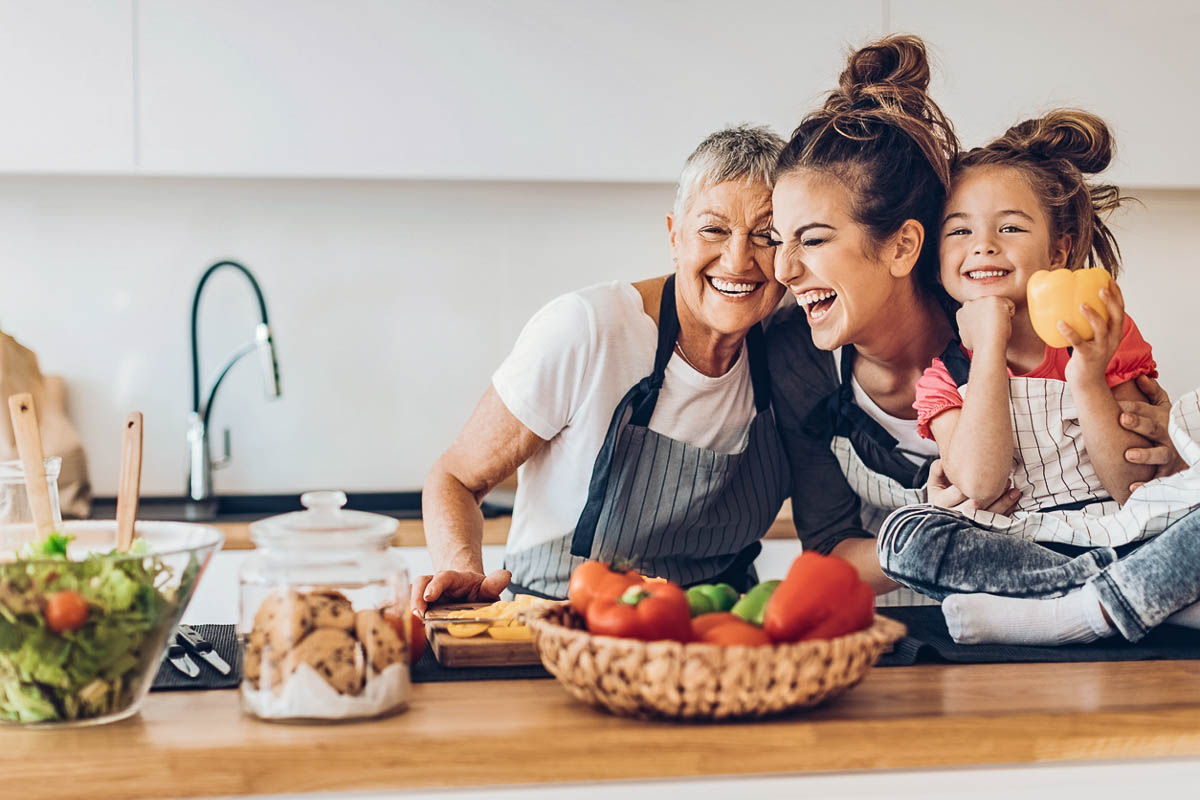 a grandma, mom and daughter in the kitchen, all smiling, the daughter holding an orange pepper. 