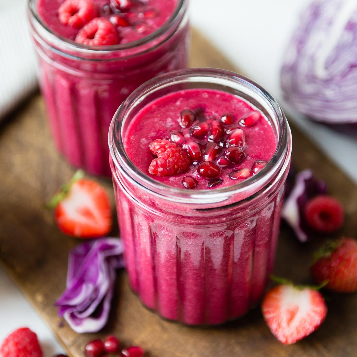 two glass jars full of pomegranate smoothies topped with pomegranate arils and raspberries, sitting on a wooden cutting board.