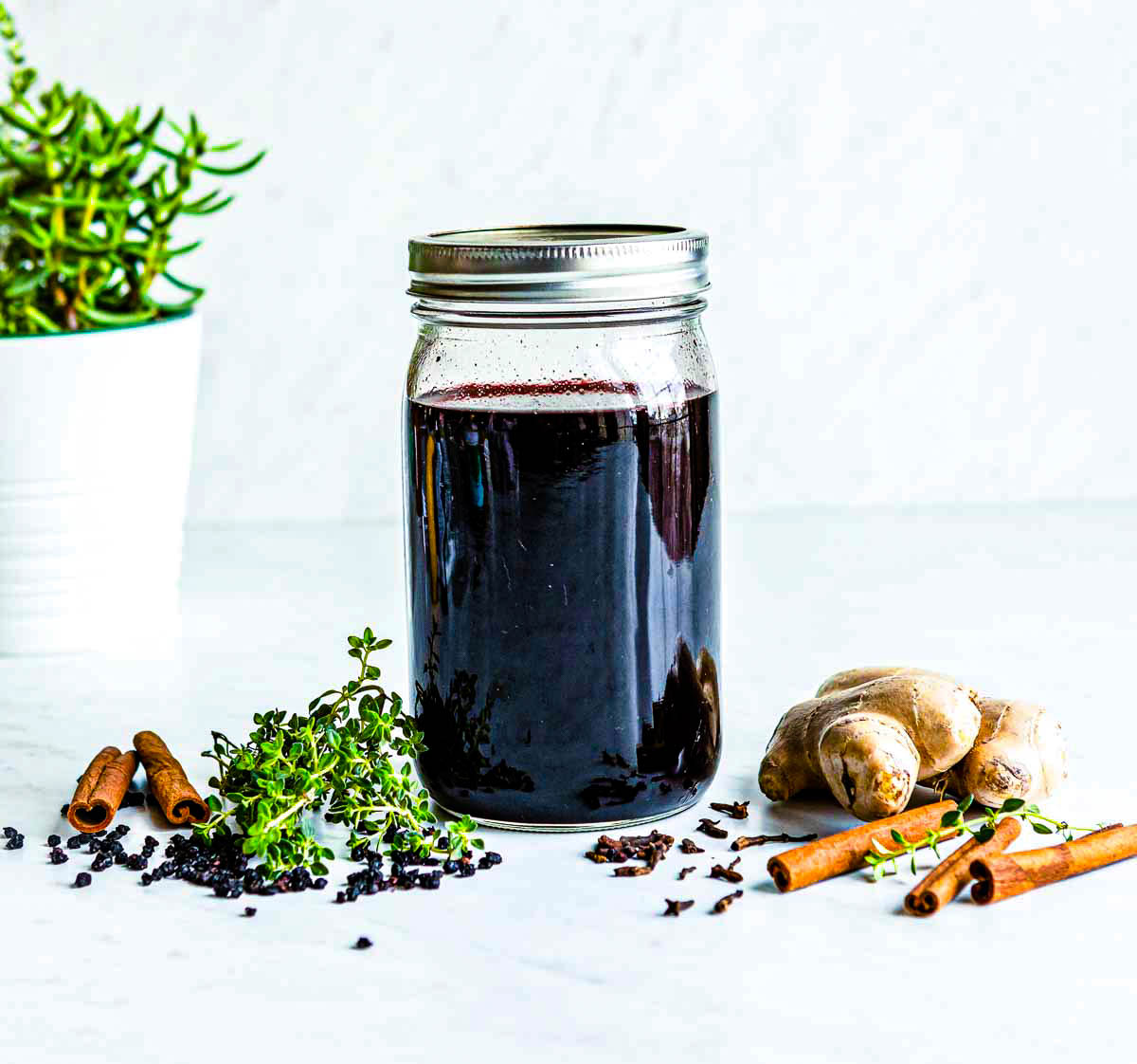 glass mason jar with elderberry syrup on a white counter top surrounded by fresh ginger, cinnamon sticks, thyme and elderberries.
