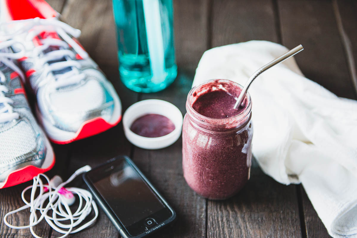 purple workout smoothie in a glass jar with a metal straw next to running shows, a phone, headphones, a water bottle and aa sweat towel. 