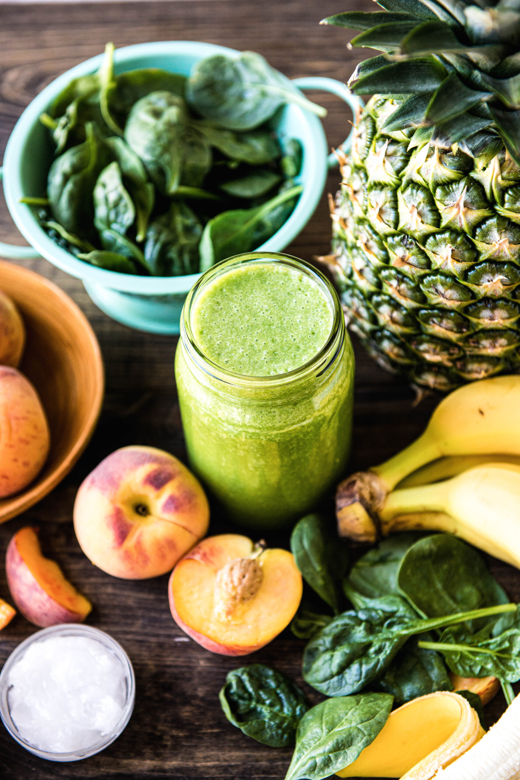 glass jar of green smoothie surrounded by fresh pineapple, banana, peach, coconut oil and spinach.