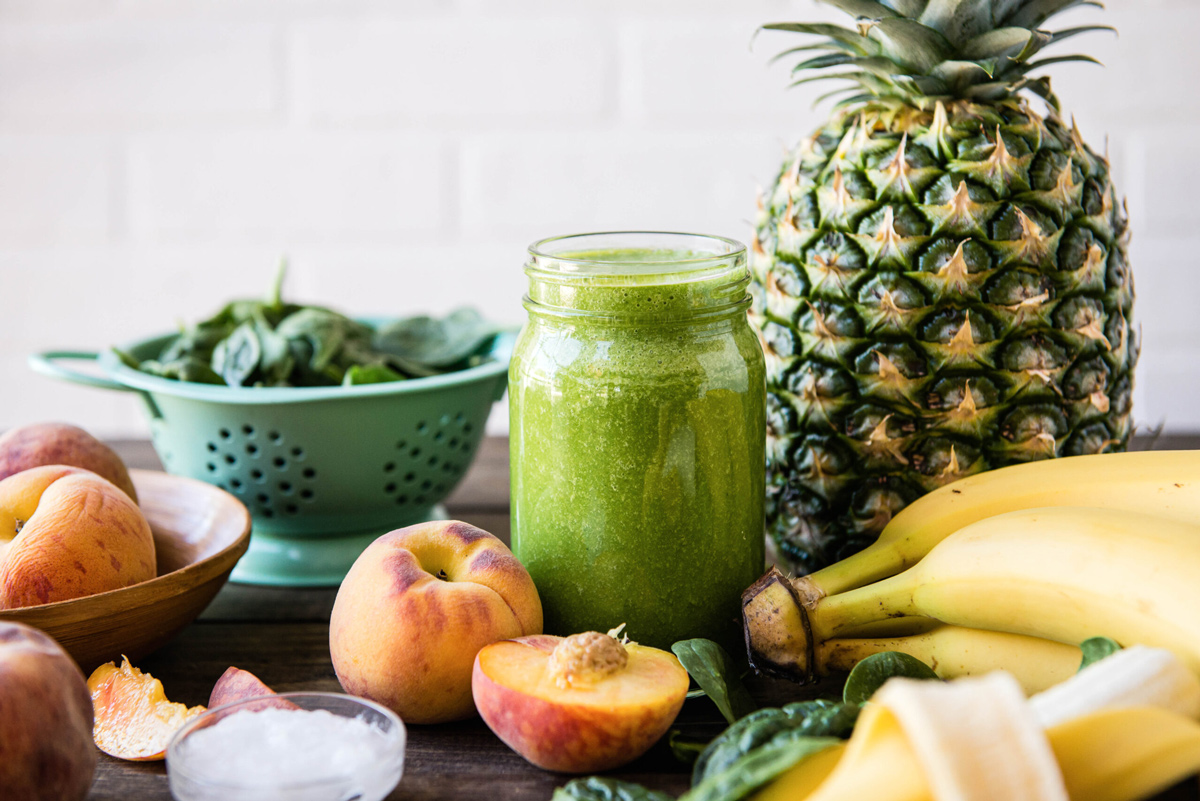Green pre-workout smoothie in a glass jar surrounded by fresh produce featuring banana, spinach, and pineapple.