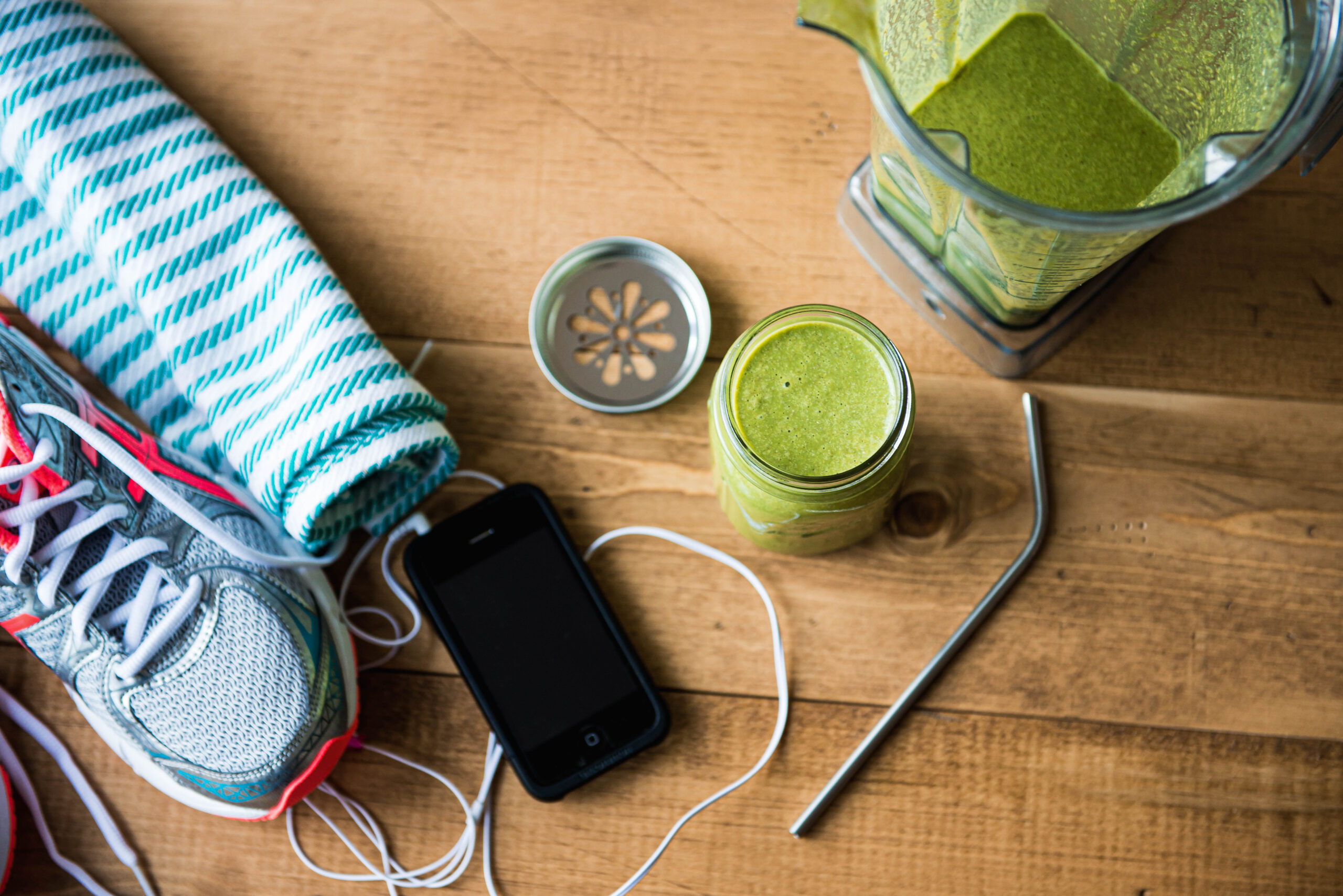 green smoothie in glass jar with flower lid and metal straw next to blender container, phone with headphones plugged in, sweat towel and running shoes.