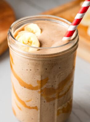 Banana smoothie in a glass with a striped straw, topped with banana slices.